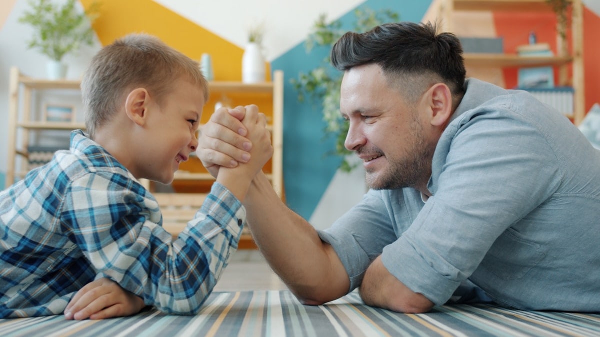 caring father and active son enjoying arm wrestling on floor at home laughing having fun together. People, sports activities and house concept.